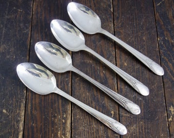 May include: Four silver spoons arranged diagonally on a dark wooden surface. The spoons have oval bowls and long, detailed handles with a decorative pattern near the end. The spoons reflect the light, creating a bright, shiny appearance. The spoons are a set of silverware.