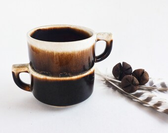 May include: Two brown and white ceramic mugs with a brown glaze stacked on top of each other. The mugs have a rounded shape and a single handle.