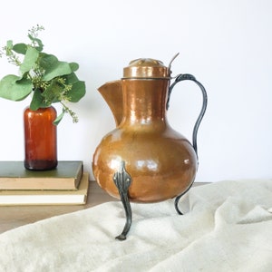 May include: A copper teapot with a hammered finish and a lid. The teapot has a curved spout and a handle. It sits on three black metal legs.