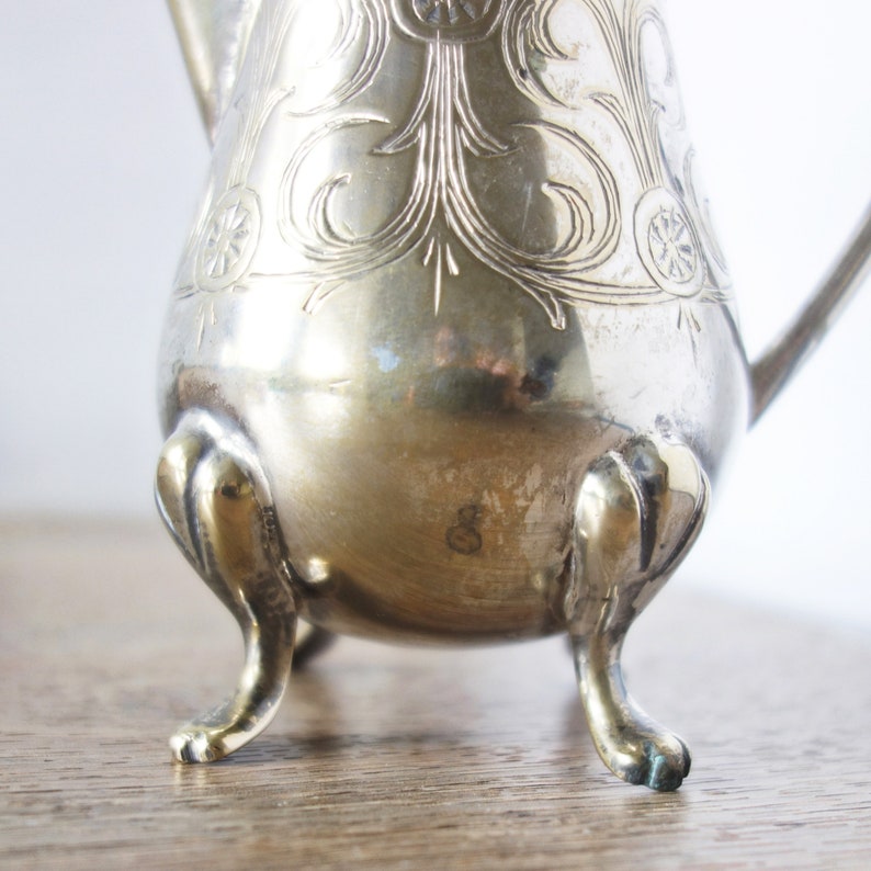 May include: A silver-colored, antique creamer with ornate floral and scrollwork detailing. The creamer has a rounded body, a spout, and a handle. It stands on three curved feet.
