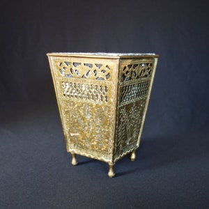 May include: A gold metal umbrella stand with an intricate, ornate design. The stand has a square base and four legs.