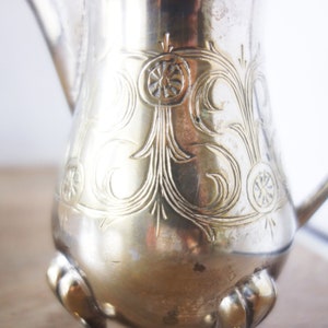 May include: A silver-colored pitcher with a decorative floral pattern and three small feet. The pitcher has a handle and a spout. The floral design is etched into the metal, adding a vintage aesthetic.