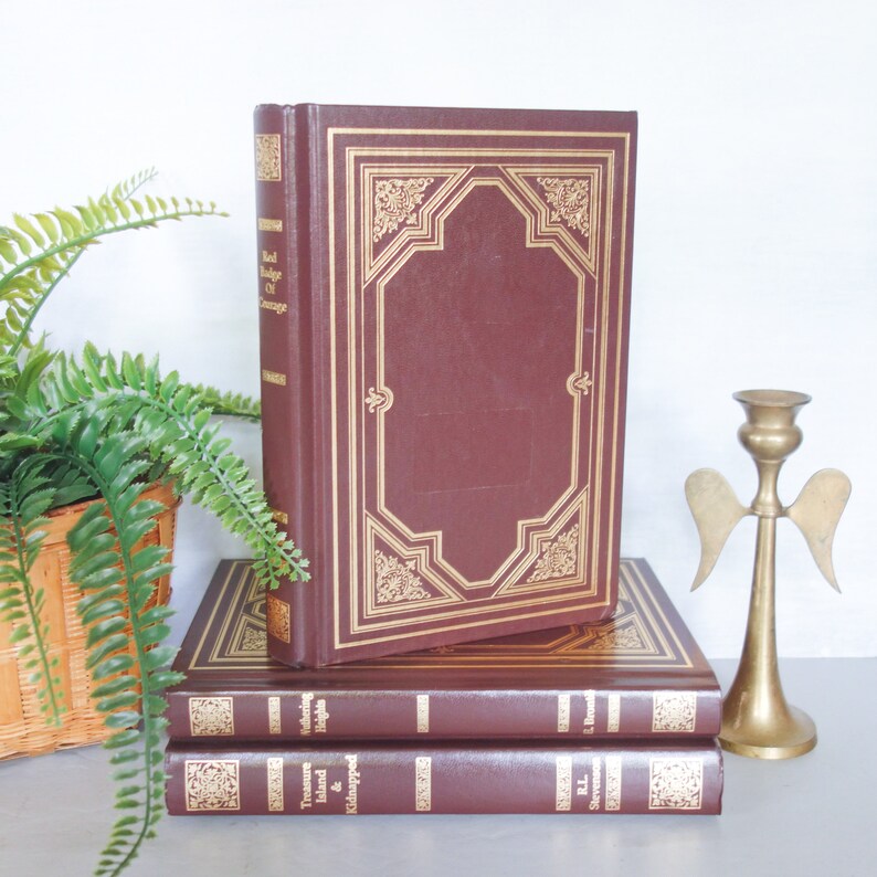 May include: A stack of three hardcover books with gold accents. The top book is titled "Red Badge of Courage" and the bottom book is titled "Treasure Island & Kidnapped".