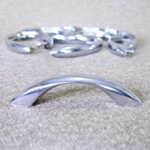 May include: A set of polished chrome cabinet pulls featuring a smooth, arched design. One pull is positioned in the foreground, while several identical pulls are arranged in a blurred, circular formation in the background, showcasing a reflective finish.