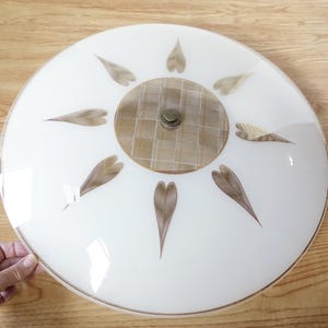 May include: A vintage white glass ceiling light fixture with a circular design. It features a central, patterned circle and is adorned with leaf-shaped accents in shades of brown. The fixture has a metal center and a brown trim.