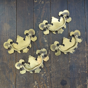 May include: Four ornate brass drawer pulls with a decorative, scroll-like design. Each pull has a curved handle and a decorative, floral-like design.