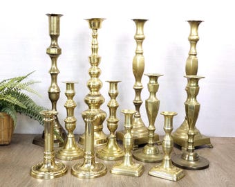 Puede incluir: Una colección de once candelabros de latón de diversas alturas y diseños, con superficies metálicas pulidas, bases circulares y cuadradas, y tallos escalonados. Los candelabros están dispuestos en grupo sobre una superficie de madera.