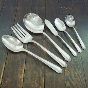 May include: A set of six silver-plated serving utensils, including a large serving spoon, a smaller serving spoon, a butter knife, a fork, and two teaspoons. The utensils are arranged on a wooden surface.