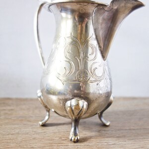 May include: A silver-colored pitcher with a curved spout and a handle. The pitcher has ornate floral engravings and rests on three claw-shaped feet. The background is a light color, and the pitcher is on a wooden surface.