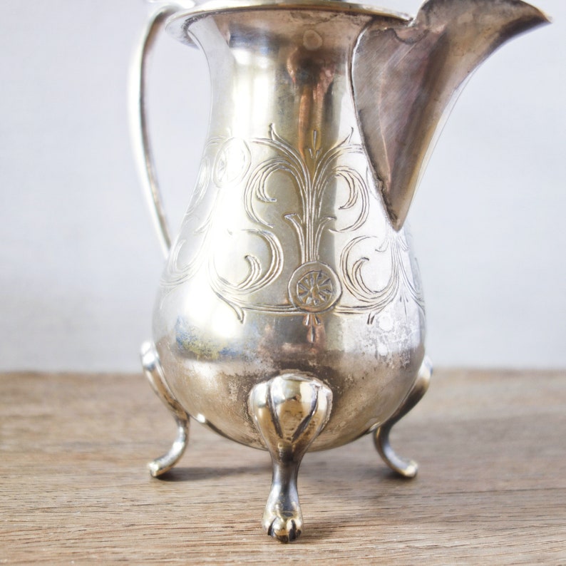 May include: A silver-colored pitcher with a curved spout and a handle. The pitcher has ornate floral engravings and rests on three claw-shaped feet. The background is a light color, and the pitcher is on a wooden surface.