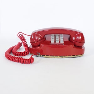 May include: A red rotary dial phone with a coiled red cord. The phone has a push-button keypad with numbers and letters.