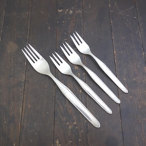 May include: Four silver forks with decorative handles are arranged diagonally on a dark wooden surface. The forks have tines and a detailed pattern on the handles. The silverware is likely for dining.