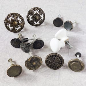 May include: A collection of various cabinet knobs including two large circular bronze-toned pieces with intricate cut-out patterns, several smaller dark textured metal knobs, a smooth white ceramic knob, and a plain circular bronze-toned knob.