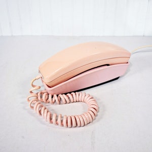 May include: A vintage pink rotary phone with a coiled cord. The phone has a simple design and is in good condition.