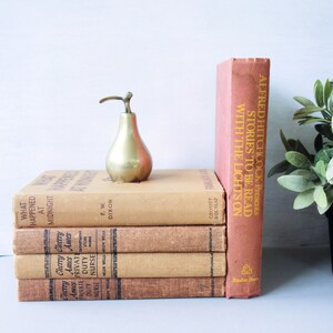 May include: A stack of vintage paperback books with brown covers and gold lettering. The books are titled "What Happened at Midnight", "Cherry Ames Private Duty Nurse", and "Alfred Hitchcock Presents Stories to Be Read With the Lights On". A small gold pear-shaped bottle stopper sits on top of the books.