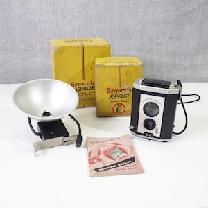 May include: A vintage Brownie Reflex camera with a flash attachment, instruction manual, and two boxes. The camera is black and silver with a chrome lens. The boxes are yellow with black text. The flash attachment is silver with a black handle.