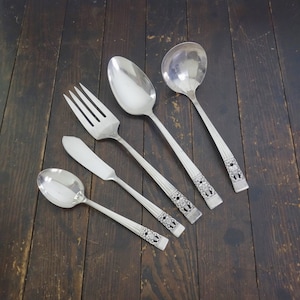 May include: Five silver-plated utensils, including a fork, butter knife, soup spoon, serving spoon, and a gravy ladle. The utensils have ornate floral designs and are arranged on a wooden surface.