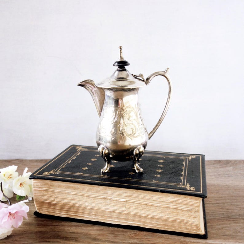 May include: A silver-colored, antique coffee pot with ornate floral engravings, a curved handle, and a black finial. The pot is displayed on a vintage book with gold detailing.
