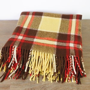 May include: A folded plaid blanket in shades of brown, red, and yellow. The blanket features a large central square and a fringe of alternating colors. The blanket is made of a soft material and is suitable for use as a throw.