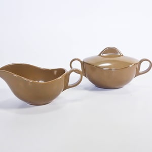 May include: A brown ceramic gravy boat and covered sugar bowl set. The gravy boat has a curved spout and a handle. The sugar bowl has a lid with a small handle and two side handles. Both pieces have a smooth, glossy finish.