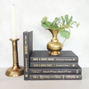 May include: A stack of five black hardcover books with gold lettering on the spines. The books are titled "Jerusalem Vigil", "Thunder for Jerusalem", "Jerusalem's Heart", "Island of Hope, Island of Tears", and "All Rivers to the Sea". The books are by Bodie and Brock Thoene. A gold candle holder with a white candle and a gold pitcher with green leaves are on a white surface in front of the books.