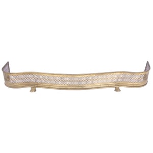 May include: A gold metal fireplace fender with a decorative, openwork design. The fender has a curved shape and two feet on each end.