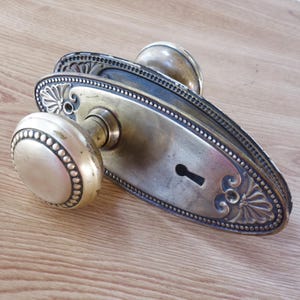 May include: An antique brass door handle set. The set includes two round doorknobs with a beaded edge and a rectangular backplate with decorative detailing. The finish is aged with a patina.