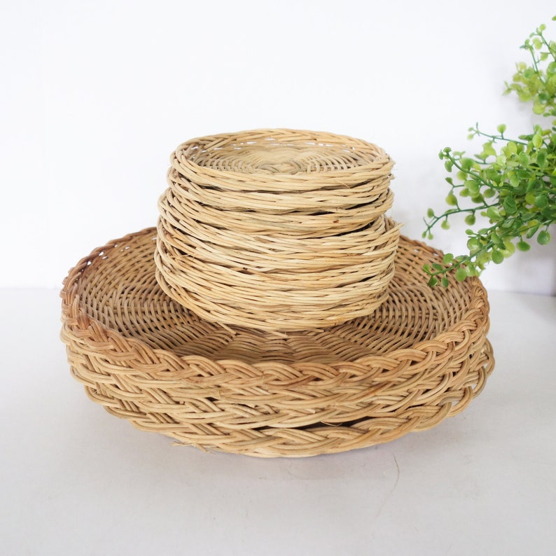 May include: A stack of round, light brown woven wicker coasters resting on a larger, circular woven wicker tray. The items feature a natural tan color and a detailed, interlaced texture throughout the circular bases and the braided edges.