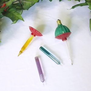May include: A collection of four vintage-style Christmas light bulbs. One bulb features a green and red dome with a brass screw base, while the others are slender, cylindrical glass tubes in yellow, teal, and purple hues with pointed tips.