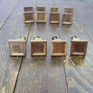 May include: Ten antique brass cabinet knobs with a square shape and a raised edge. The knobs have a brushed finish and are arranged in two rows on a wooden surface.