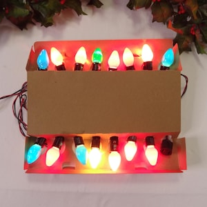 May include: A set of vintage Christmas lights in a cardboard box. The lights are various colors, including blue, green, and white. The box is open, revealing the bulbs and their wiring. The lights are arranged in two rows.