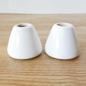 May include: Two white ceramic bud vases with a rounded, conical shape. The vases have a glossy finish and a small opening at the top. They are displayed on a light-colored wooden surface.