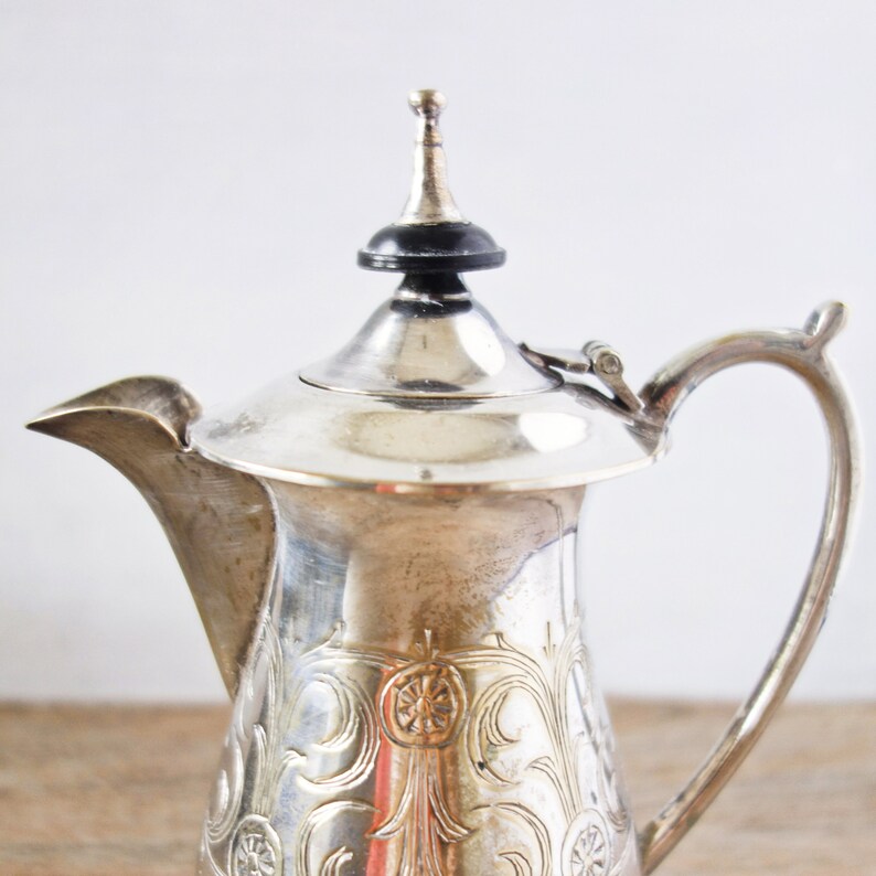 May include: A silver-colored tea or coffee pot with a decorative floral pattern. The pot has a curved handle, a spout, and a lid with a black finial. The pot is made of metal and appears to be vintage.