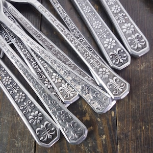 May include: A set of silver-toned flatware with a decorative floral and fleur-de-lis pattern.