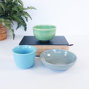 May include: Three ceramic bowls in shades of green, blue, and gray. The green bowl sits atop a dark book, while the blue and gray bowls are on a white surface. The bowls vary in shape and size, showcasing a minimalist design.