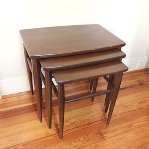 May include: A set of three nesting tables in varying sizes. The tables are made of dark brown wood with a rectangular top and tapered legs. The tables are arranged in a stacked configuration, with the largest table at the bottom.