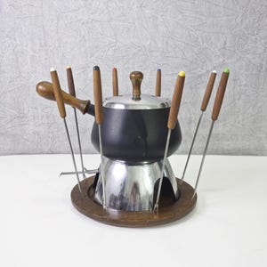 May include: A black fondue pot with a wooden base and eight metal fondue forks with wooden handles. The forks have colorful plastic tips.