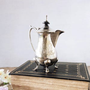 May include: A silver-colored, antique coffee pot with ornate floral engravings. The pot has a curved handle, a spout, and a decorative lid. It stands on three small feet and is placed on a vintage book with gold-edged pages.