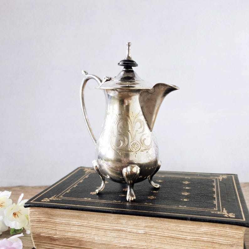 May include: A silver-colored, antique coffee pot with ornate floral engravings. The pot has a curved handle, a spout, and a decorative lid. It stands on three small feet and is placed on a vintage book with gold-edged pages.