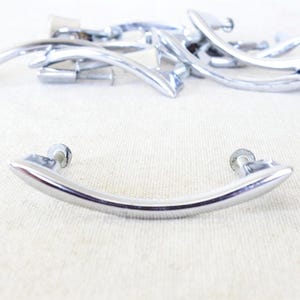 May include: A collection of polished chrome drawer pulls with a curved, arching design. Each handle features two mounting points with visible metal screws extending from the back, showcasing a smooth, reflective metallic finish throughout.
