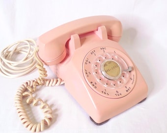 May include: A pastel pink rotary telephone featuring a circular dial with black numbers and letters. The handset rests on the base, connected by a matching coiled cord. The centre of the dial displays the phone number 686-0653 in black text.