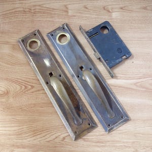 May include: Antique door hardware set, including two long rectangular plates with handles and a separate latch mechanism. The plates are a tarnished brass color, and the latch is dark blue. The set is likely from an older home.