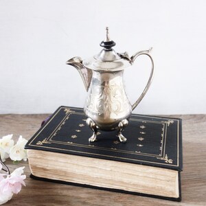 May include: A silver-colored antique teapot with ornate floral engravings, a curved handle, and a black finial. The teapot is displayed on a large, antique book with a black cover and gold detailing.