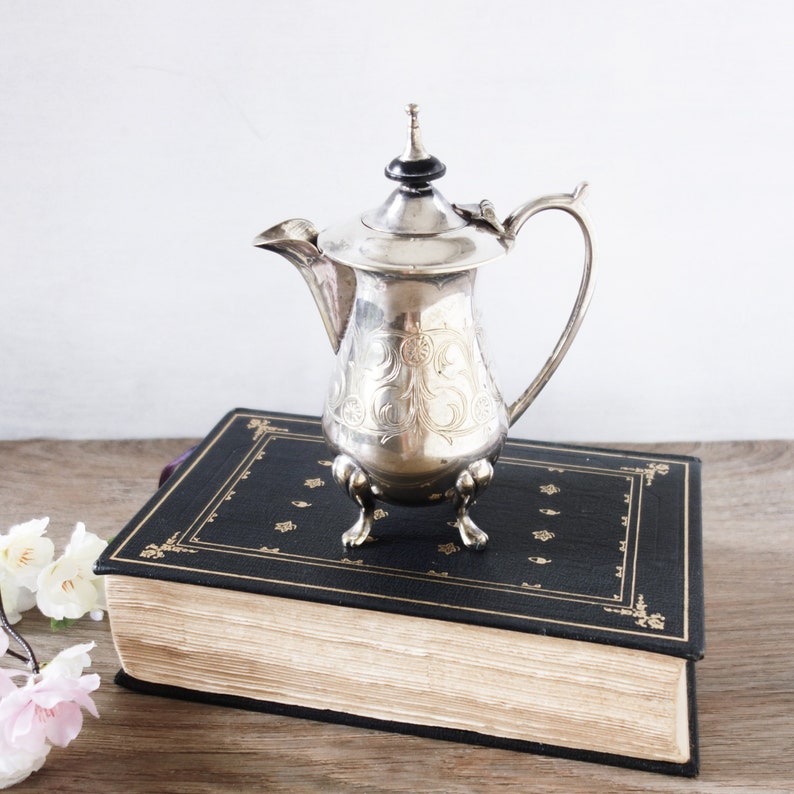 May include: A silver-colored antique teapot with ornate floral engravings, a curved handle, and a black finial. The teapot is displayed on a large, antique book with a black cover and gold detailing.