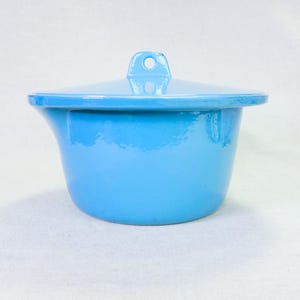 May include: A light blue enamelware pot with a matching lid. The pot has a rounded shape with a flared rim and a small handle on the lid. The pot is a solid, glossy light blue color.