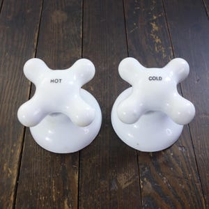 May include: Two white porcelain faucet handles with a cross-shaped design. One handle is labeled "HOT" and the other "COLD" in black text. The handles are mounted on round bases.