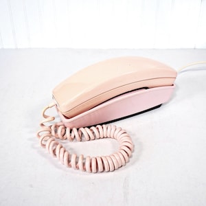May include: A vintage-style landline telephone in a soft pastel pink hue. The device features a smooth, curved handset resting on a matching base, connected by a coiled, flexible cord that extends outward from the side of the unit.