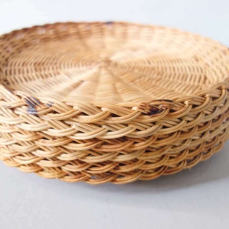May include: A round, shallow tray woven from light tan natural reeds. The tray features a tight, circular weave pattern on the base and a decorative, braided border along the raised edge, showcasing variations in the natural color of the reeds.