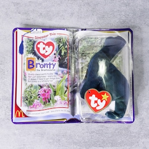 May include: A new, unopened Ty Beanie Baby Bronty the Brontosaurus plush toy in its original packaging. The packaging includes a purple border, the McDonald's logo, and the text "Dinosaur Trio". The plush toy is dark blue.