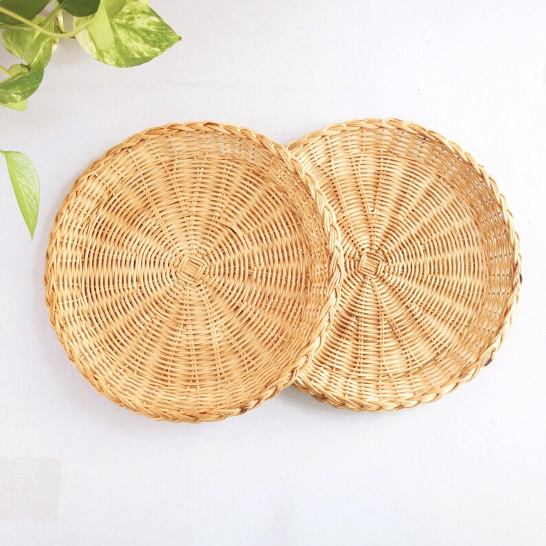 May include: Two round, shallow woven baskets made of light tan natural reed. Each basket features a tight, circular radial weave pattern with a braided rim edge. The baskets are positioned side-by-side, showcasing a consistent light brown color.